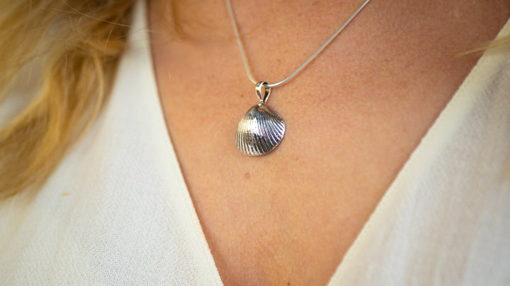 A close-up of a person wearing the Wildlife Pendant - Cockle Shell With Chain from Gallardo & Blaine Designs. The person is dressed in a white top, and the focus is on the intricate cockleshell pendant resting against their skin.