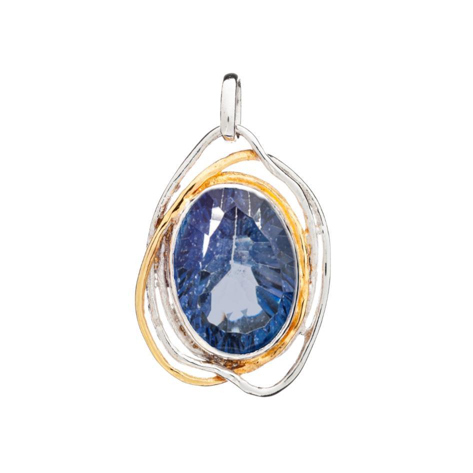 A blue gemstone pendant with a faceted oval sapphire encased in an intricate metal frame featuring silver and gold tones. With its medieval design, the Cosmos Pendant Large in various gemstones by Gallardo & Blaine Designs has a loop at the top for a chain attachment, making it a true statement gemstone pendant.