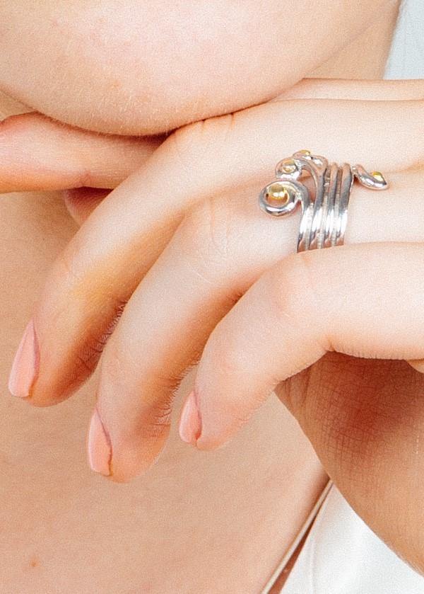 Close-up of a person's hand resting on their chin, showcasing the **Crown Ring** by **Gallardo & Blaine Designs**, an adjustable silver ring with decorative swirls and small yellow gemstones. The person has light pink nail polish and light skin.