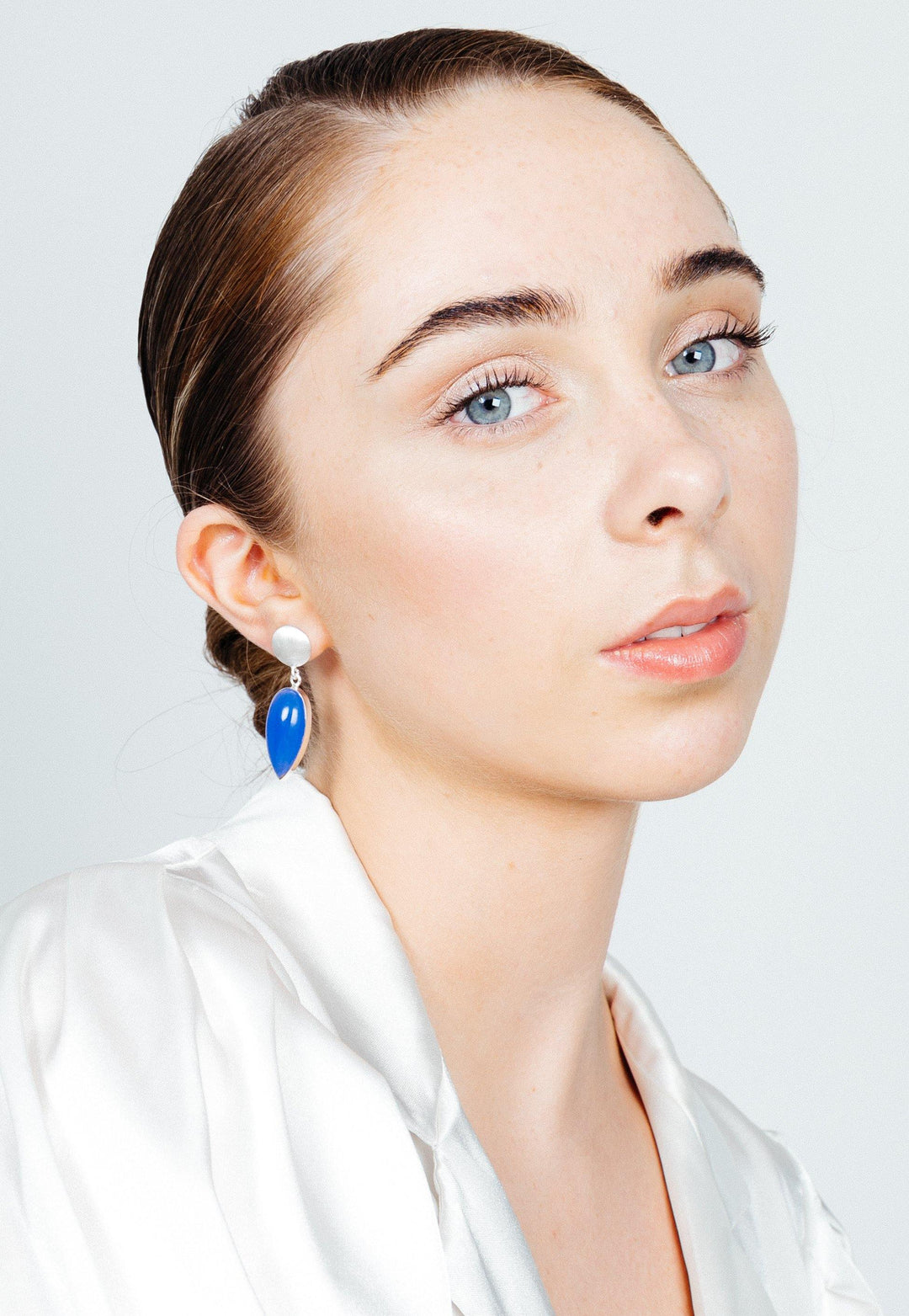 A young person with fair skin, light blue eyes, and brown hair pulled back looks into the camera. They wear a white satin blouse and Gallardo & Blaine Designs' Daira Earrings in sterling silver, featuring a white bead and a blue, elongated teardrop-shaped charm. The background is light gray.