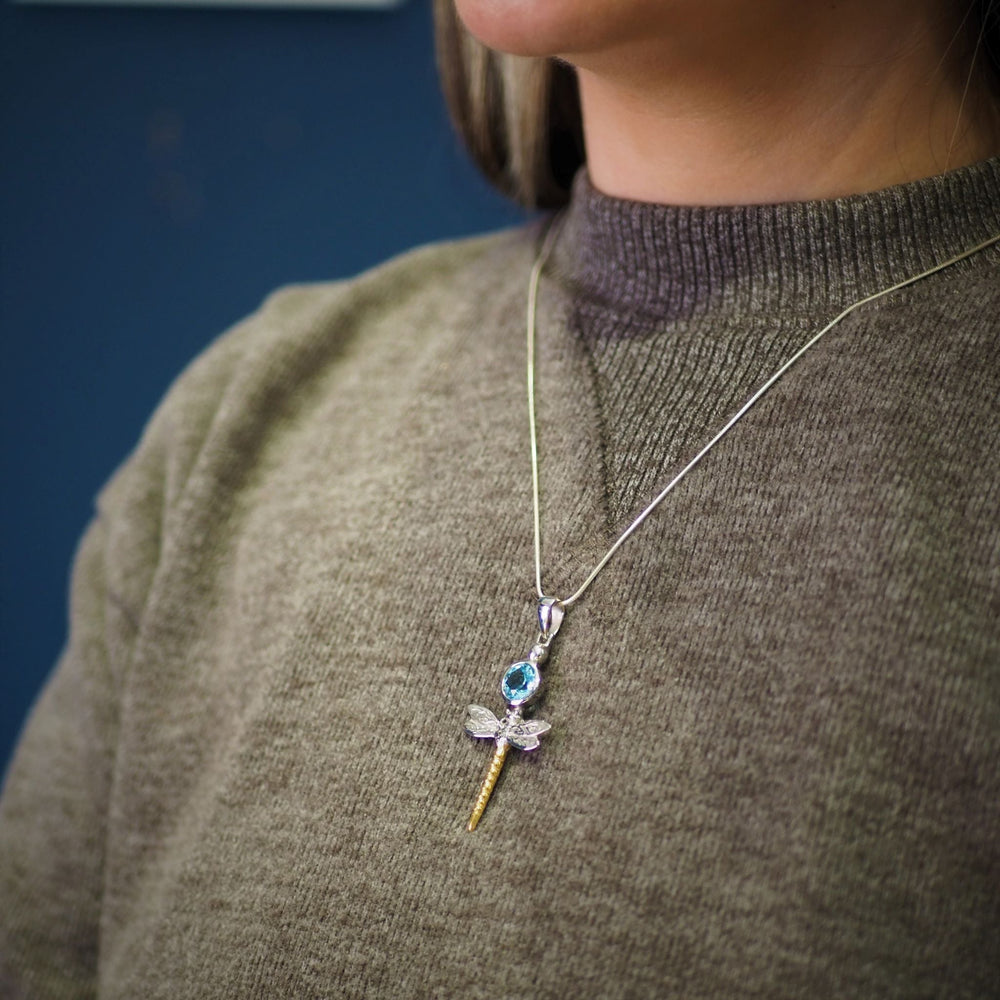 A person wearing a green sweater is shown from the shoulders up. They're adorned with a Damsel Pendant in various gemstones, a sterling silver necklace shaped like a sword or dagger, showcasing a blue gemstone in the hilt and small wings near the blade by Gallardo & Blaine Designs. The background is a dark blue.