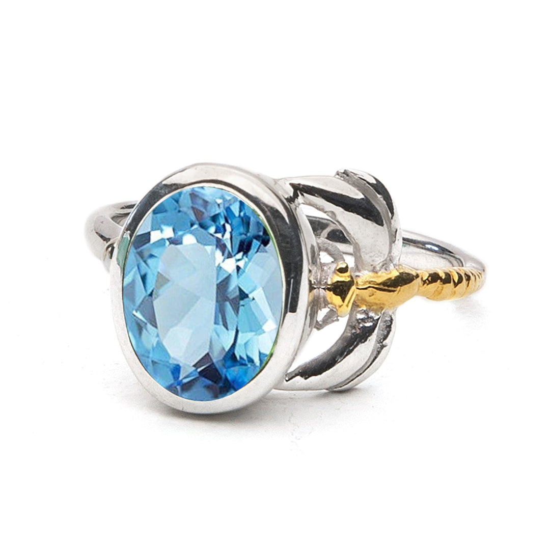 A timeless piece, the Daydream Ring in various gemstones by Gallardo & Blaine Designs features a large, oval-shaped blue gemstone in the center. The gemstone is encased in a polished silver setting and flanked by delicate metalwork resembling leaves or petals, with a small gold accent on one side.