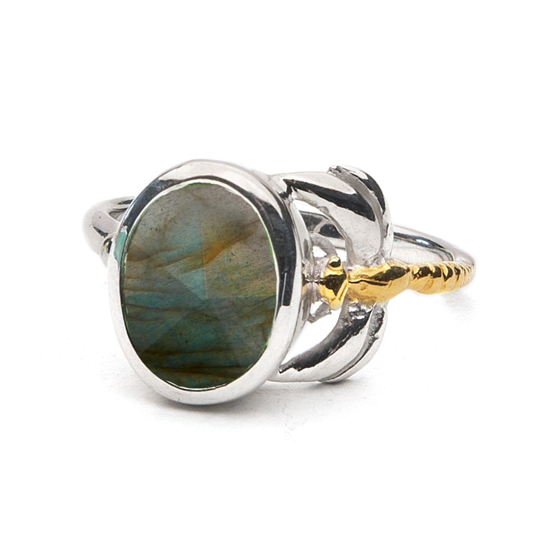 A close-up image of the Daydream Ring in various gemstones by Gallardo & Blaine Designs featuring an oval-shaped labradorite gemstone set in a silver band with gold accents designed to resemble leaves or petals. This timeless piece displays a range of blue and green colors, embodying delicate and dainty craftsmanship.