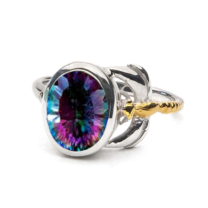 A Gallardo & Blaine Designs Daydream Ring in various gemstones featuring a multicolored, oval-shaped gemstone in the center with shades of purple, blue, and green. The delicate and dainty design includes intricate golden anchor details on either side of the gemstone, adding a nautical element to this timeless piece.