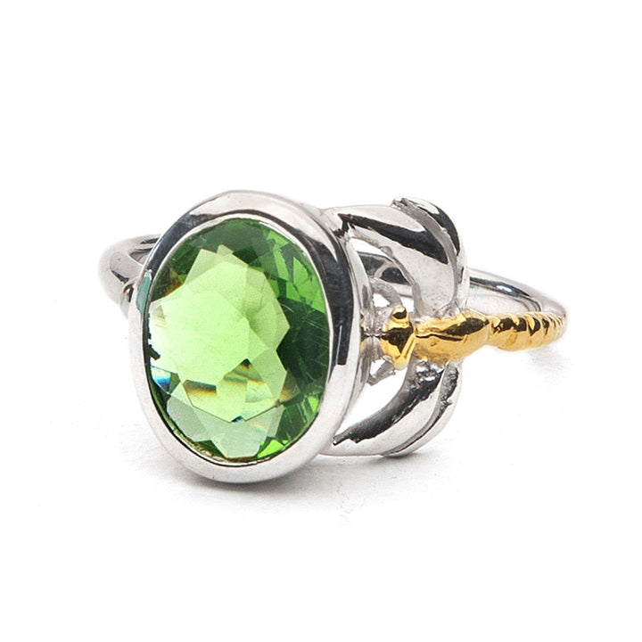 The Daydream Ring in various gemstones by Gallardo & Blaine Designs features a large, round, faceted green gemstone in the center. The setting has a unique design that includes gold accent details on the sides, resembling small leaves or petals. The smooth and shiny band adds to the charm of this delicate and dainty piece.