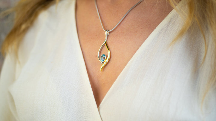 Close-up of a person wearing a sterling silver necklace with a teardrop-shaped pendant. The pendant, known as the Desert Star Pendant in various gemstones by Gallardo & Blaine Designs, features a small blue gemstone at the center and is worn over a white V-neck top. The background is blurred.