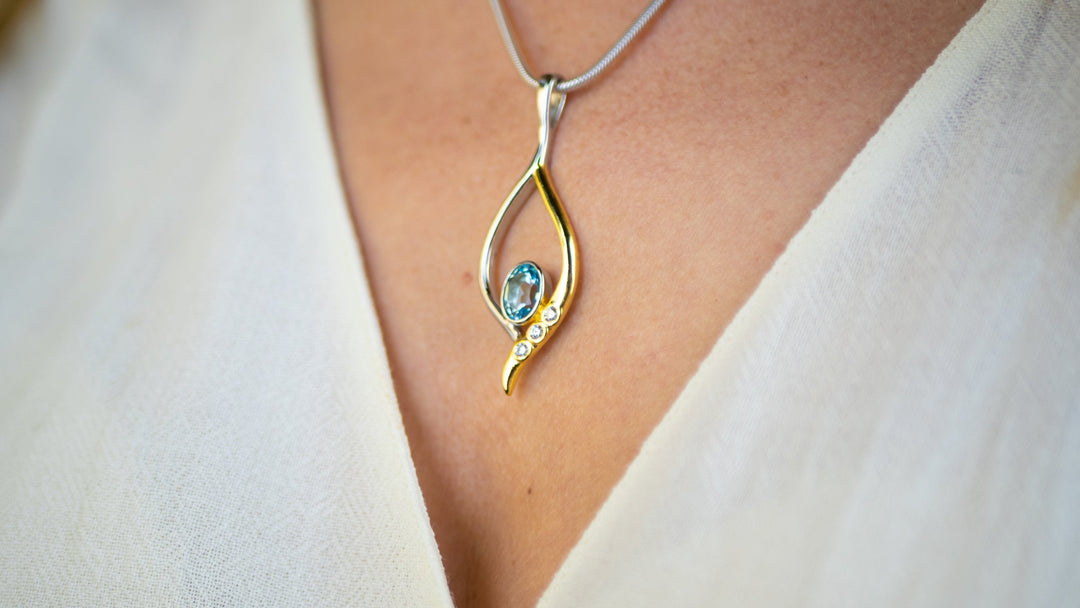 A close-up image of a person wearing a white garment, showcasing a delicate Desert Star Pendant in various gemstones by Gallardo & Blaine Designs. The pendant, crafted in gold vermeil and sterling silver, features a small blue gemstone at its center, adorned with a few smaller clear stones accenting its lower half.