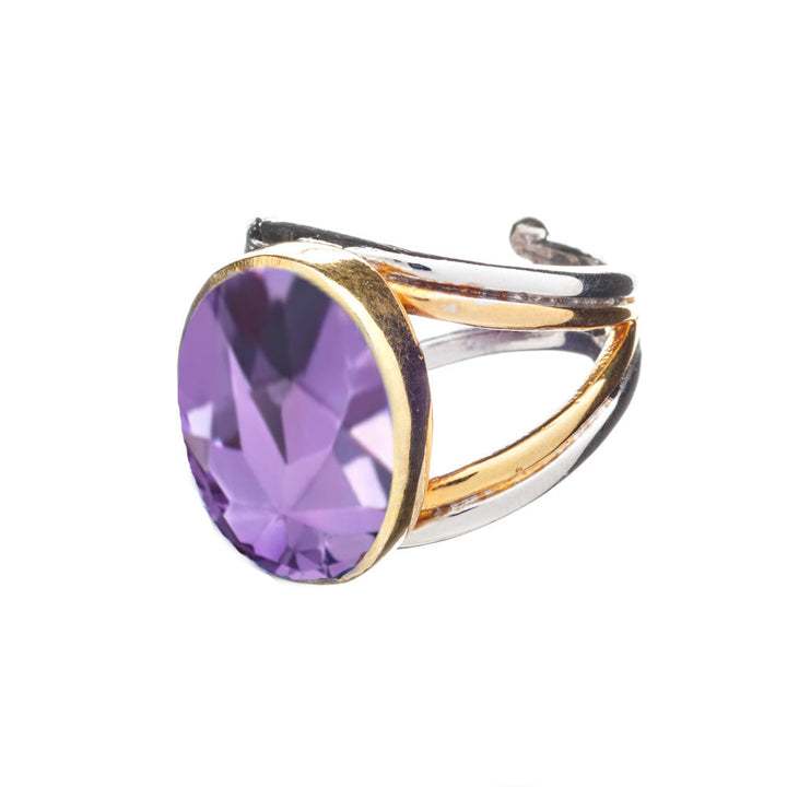 A chunky statement ring featuring a large oval-shaped purple gemstone set in a golden bezel. The sterling silver band has an open, adjustable design, forming an asymmetrical stylish appearance. The faceted gemstone catches light beautifully, displaying various shades of purple. This is the Dewberry Ring in various gemstones by Gallardo & Blaine Designs.