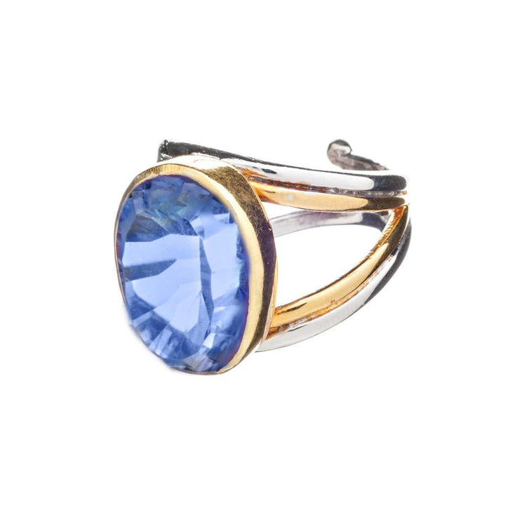 A close-up image of a chunky statement ring featuring a large, oval-shaped blue gemstone set in a band that combines sterling silver and gold elements. The band has a unique, open design, adding an elegant touch to the overall appearance of the Dewberry Ring in various gemstones by Gallardo & Blaine Designs.