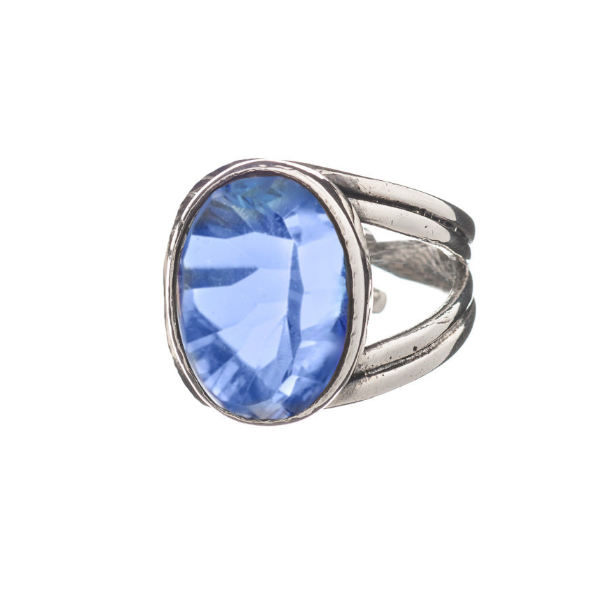 A close-up image of the Dewberry Ring in various gemstones featuring a large oval-shaped blue gemstone by Gallardo & Blaine Designs. The sterling silver band is textured and splits into two near the faceted stone. Set against a white background, the ring's bold design and vibrant color truly stand out.