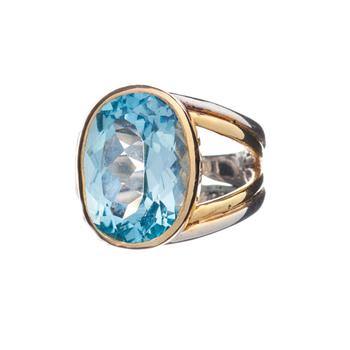 A chunky sterling silver ring featuring a large, oval-shaped blue gemstone in the center, framed by a gold bezel setting. The wide band splits into two parts on each side of the stone, enhancing its decorative appeal. The adjustable Dewberry Ring in various gemstones by Gallardo & Blaine Designs sits isolated against a white background.