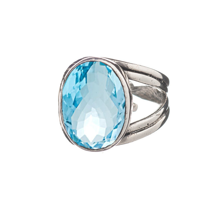 A Gallardo & Blaine Designs Dewberry Ring featuring a large, oval-shaped, faceted blue gemstone set in the center. The band appears to split into two near the gemstone, enhancing its elegant design. This chunky statement ring stands out against the white background, commanding attention with its bold presence.