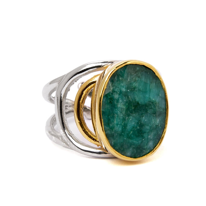 The Dewberry Ring in various gemstones by Gallardo & Blaine Designs is a chunky statement ring featuring a large, oval-shaped emerald with a textured surface, set in a gold bezel. The band is made of sterling silver and has a unique, open design with intertwining loops. The combination of metals and the bold gemstone creates a striking, elegant piece of jewelry.