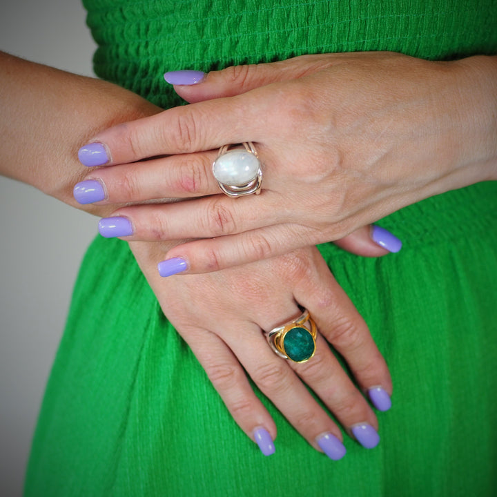 A person with light skin wearing a green dress has their hands crossed. They have lavender nail polish and are wearing two chunky statement rings: one Dewberry Ring in various gemstones by Gallardo & Blaine Designs with a large white stone on the middle finger and another Dewberry Ring in various gemstones by Gallardo & Blaine Designs with a large green stone on the middle finger of the other hand.