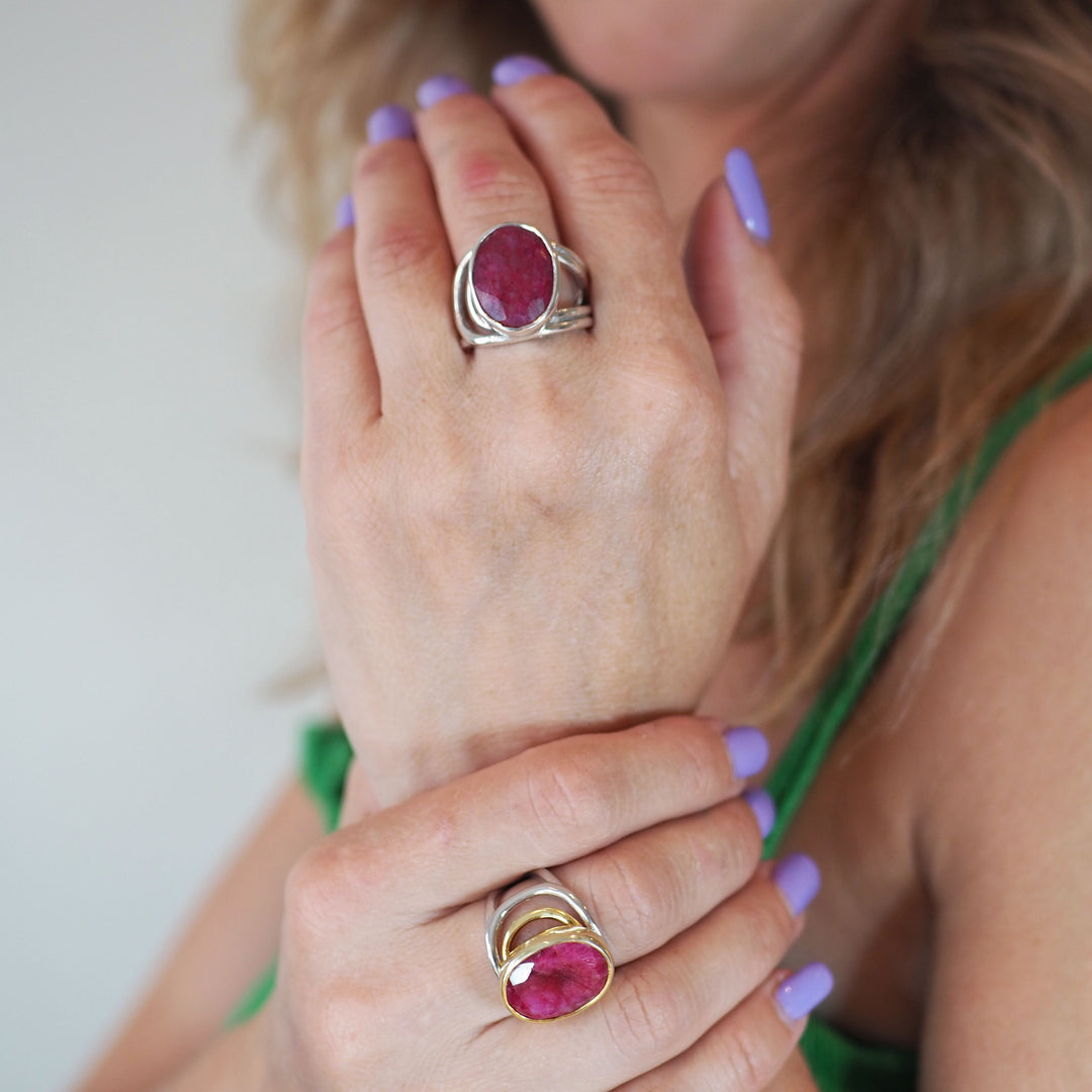 A person is showing two chunky Dewberry Rings in various gemstones by Gallardo & Blaine Designs on their fingers. The nails are painted lavender, and they are wearing a green sleeveless top. The background is blurred.