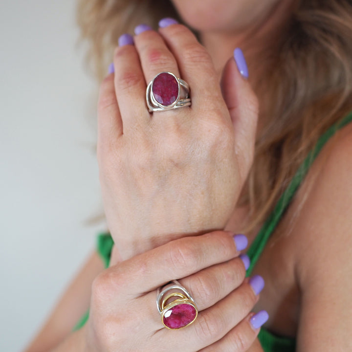 A person is showing two chunky Dewberry Rings in various gemstones by Gallardo & Blaine Designs on their fingers. The nails are painted lavender, and they are wearing a green sleeveless top. The background is blurred.