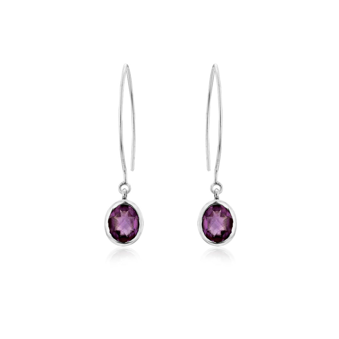 A pair of elegant sterling silver drop earrings featuring oval-shaped purple gemstones. Each earring has a thin, sleek hook leading to a bezel setting that holds the amethyst-like stones. The reflective, faceted gemstones add a touch of sophistication to these Dew Drop Earrings in various gemstones by Gallardo & Blaine Designs.