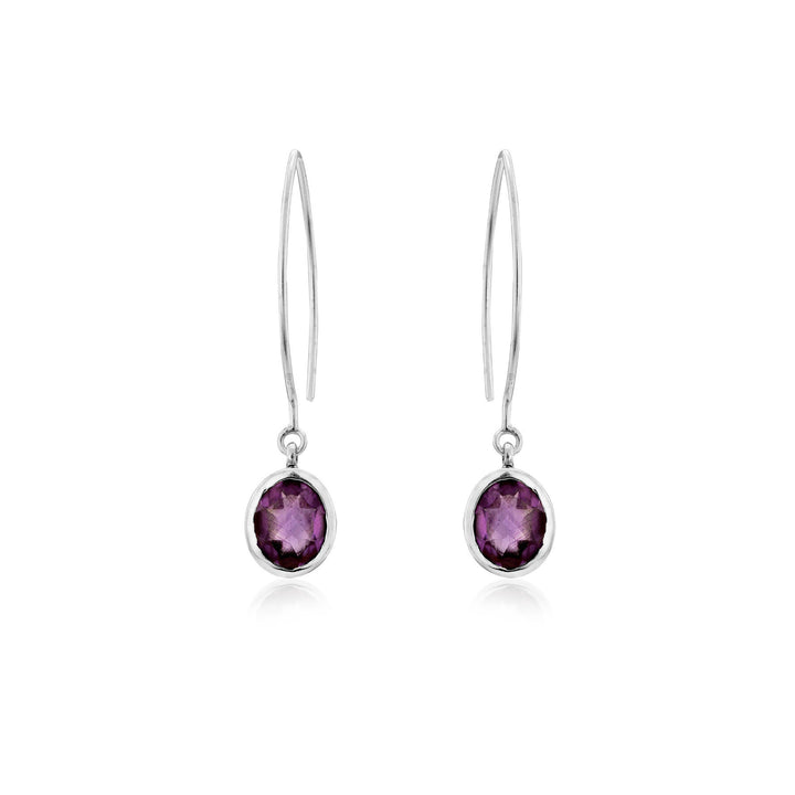 A pair of elegant sterling silver drop earrings featuring oval-shaped purple gemstones. Each earring has a thin, sleek hook leading to a bezel setting that holds the amethyst-like stones. The reflective, faceted gemstones add a touch of sophistication to these Dew Drop Earrings in various gemstones by Gallardo & Blaine Designs.