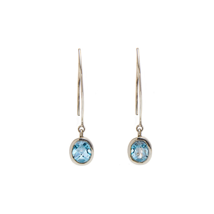 A pair of elegant Dew Drop Earrings in various gemstones featuring oval-shaped blue gemstones by Gallardo & Blaine Designs. The stones are set in a simple, silver-toned bezel setting, hanging from sleek, curved hooks. The minimalistic design highlights the sparkling blue gems.