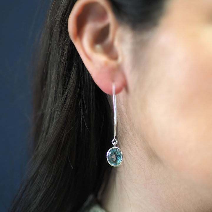 Close-up of a person wearing a long, elegant hoop design earring featuring a round, greenish-blue gemstone. The person's ear and part of their dark hair are visible against a blurred dark background, highlighting the sterling silver elegance of the Dew Drop Earrings in various gemstones by Gallardo & Blaine Designs.