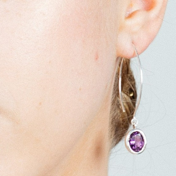 Close-up of the side of a person's face with a focus on their ear, adorned by elegant Dew Drop Earrings in various gemstones from Gallardo & Blaine Designs. The background is a neutral, light grey color.