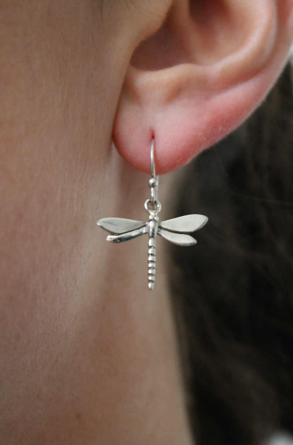 A close-up of an ear wearing Gallardo & Blaine Designs’ Wildlife Dragonfly Dangle Earrings in silver, featuring detailed wings and a segmented body. This symbolic piece stands out against the blurred background.