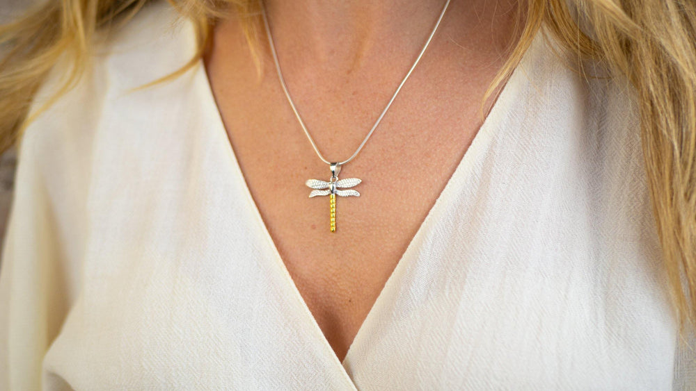 Close-up of a woman's neck adorned with a delicate silver snake chain featuring the Wildlife Pendant - Large Dragonfly With Chain from Gallardo & Blaine Designs. The dragonfly has detailed wings and a gold-toned body. She is dressed in a light beige, V-neck top.