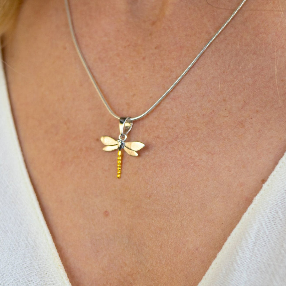 Close-up of a necklace from the Wildlife Pendant - Small Dragonfly With Chain by Gallardo & Blaine Designs, featuring a dragonfly pendant made of sterling silver and gold accents, worn by a person dressed in a light beige top. The background is softly blurred.