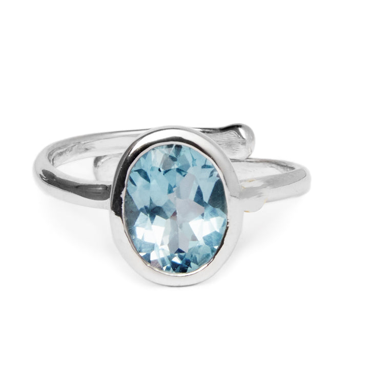 The Dream Ring in various gemstones by Gallardo & Blaine Designs is a sterling silver, adjustable ring featuring an oval-shaped blue gemstone set in a simple band. The gemstone has facets that catch the light, creating subtle highlights and reflections. The minimalist design is complemented by the smooth, polished finish of the band.