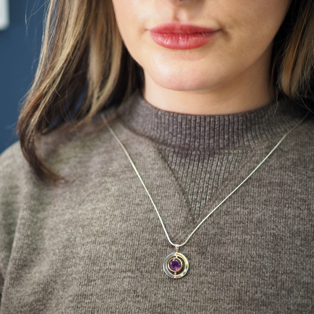 A person wearing a gray sweater and a silver necklace with a circular pendant that features a small purple gemstone. The focus is on the Elodie Pendant in silver gold & various gemstones by Gallardo & Blaine Designs, highlighting its intricate design and the gemstone's color. The background is blurred.