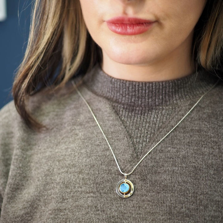 A close-up of a person wearing a brown sweater and an Elodie Pendant in silver gold & various gemstones from Gallardo & Blaine Designs. The sterling silver pendant features a blue gemstone in the center. The person has light-colored hair with darker streaks and is indoors, against a blurred blue background.