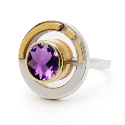 The Elodie Ring in Silver Gold & various gemstones by Gallardo & Blaine Designs showcases a round silver band adorned with a gold accent and a central purple gemstone. Blending vintage style glamour with its intricate, modern design, circular elements beautifully frame the captivating gemstone.