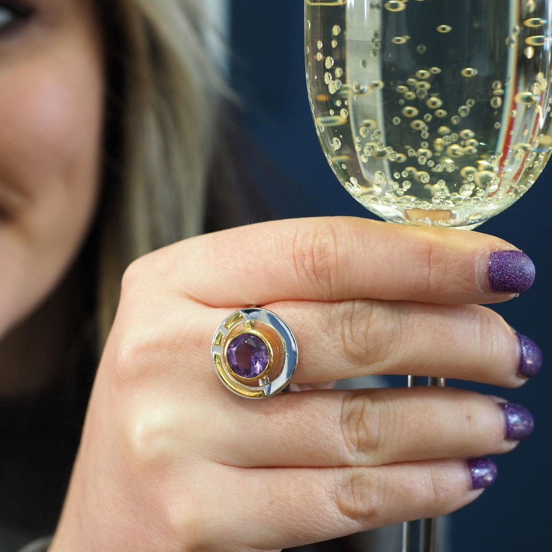 A person with purple glitter nail polish holds a glass of bubbly champagne. They are wearing the Gallardo & Blaine Designs Elodie Ring in Silver Gold & various gemstones, which features a large purple gemstone set in a gold and silver design. Part of their face is visible, with a smile showing on the left edge of the image, radiating vintage style glamour.