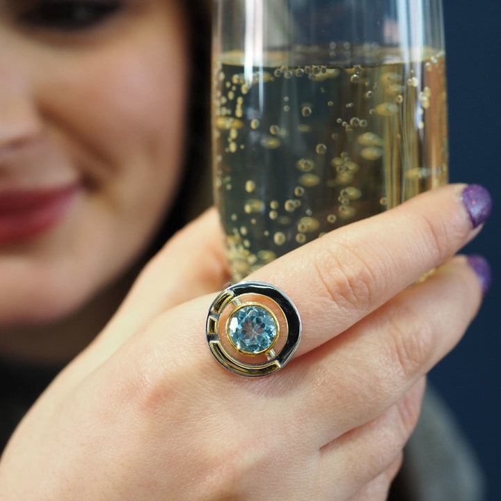 Close-up of a person holding a glass of sparkling beverage. The focus is on their hand, adorned with the Gallardo & Blaine Designs Elodie Ring in Silver Gold & various gemstones, exuding vintage style glamour. The person's face is slightly blurred in the background.