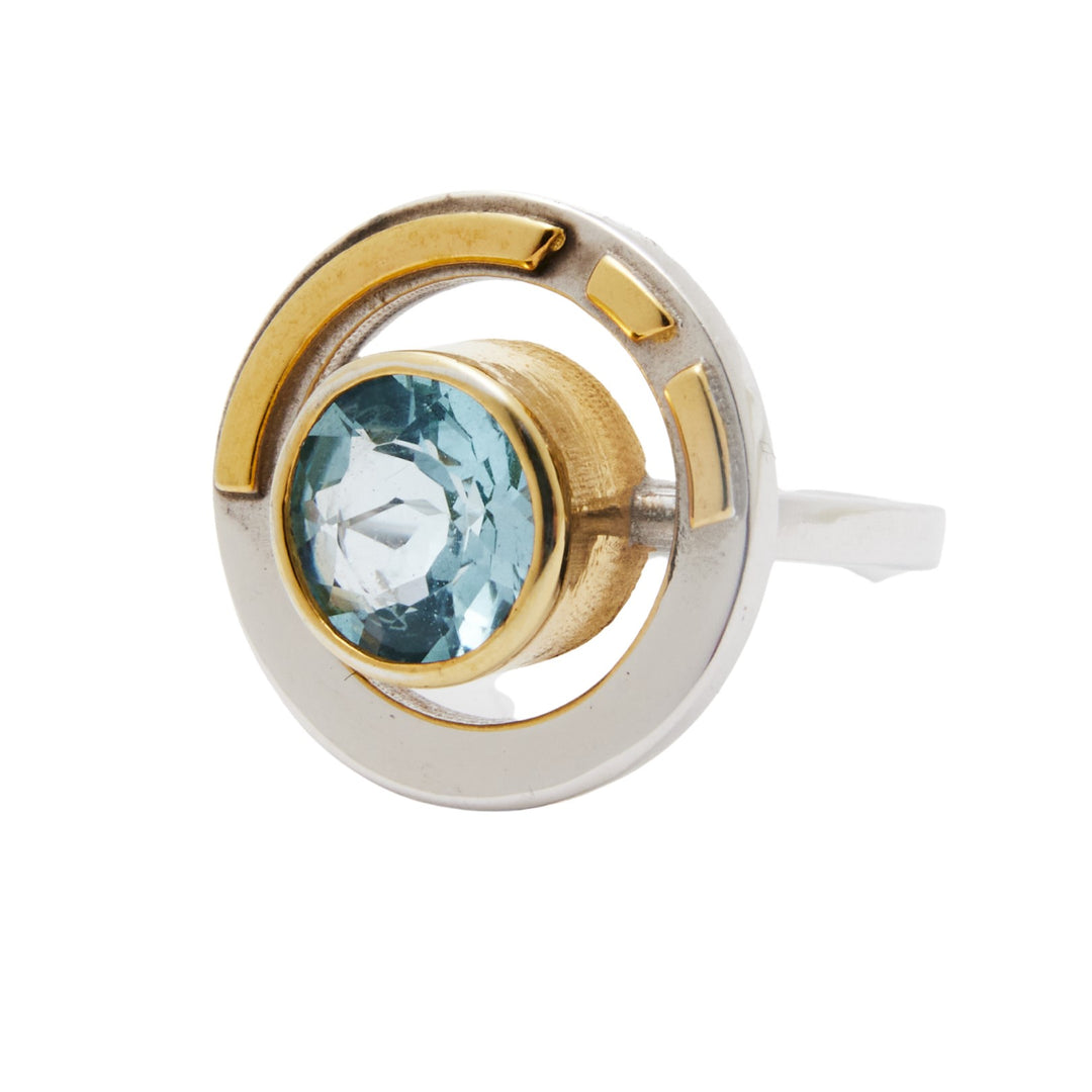 Gallardo & Blaine Designs presents the Adjustable Elodie Ring in Silver Gold, an Art Deco masterpiece with vintage glamour. Featuring a central light blue gemstone in a gold-toned bezel, it is adorned with gold accents that enhance its circular design; a timeless addition to any collection.