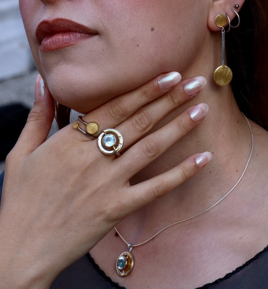 A woman with light pink nails touches her neck, wearing a Gallardo & Blaine Designs Elodie Necklace in silver and gold with gemstones, paired with a matching ring and modern gold and silver drop earring.