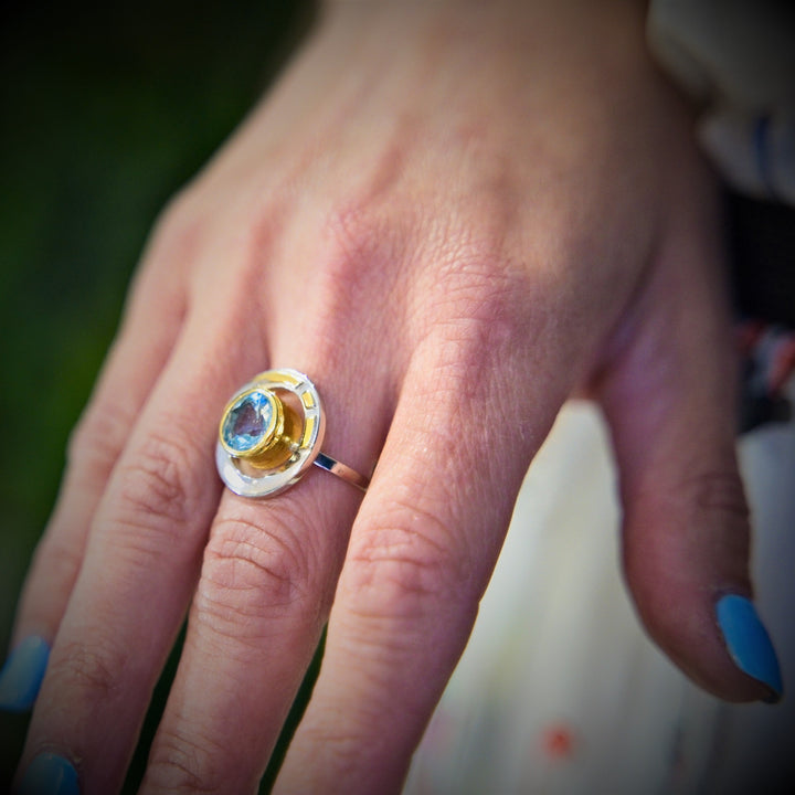 A close-up image of a hand with light blue nail polish wearing the Elodie Ring in Silver Gold & various gemstones by Gallardo & Blaine Designs. The ring features a yellow and silver band with a round blue gemstone at its center, exuding vintage style glamour. The background is slightly blurred, highlighting the hand and showcasing the ring's intricate details.