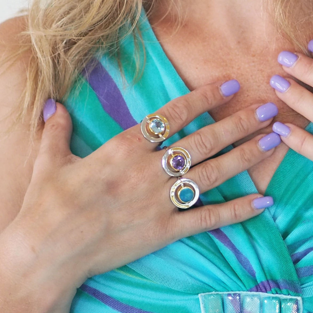 Wearing Gallardo & Blaine's Adjustable Elodie Ring in Silver Gold with vibrant gemstones, a person with lavender nail polish elegantly displays three exquisite rings, including one with vintage flair, perfectly complementing their turquoise and purple patterned top.