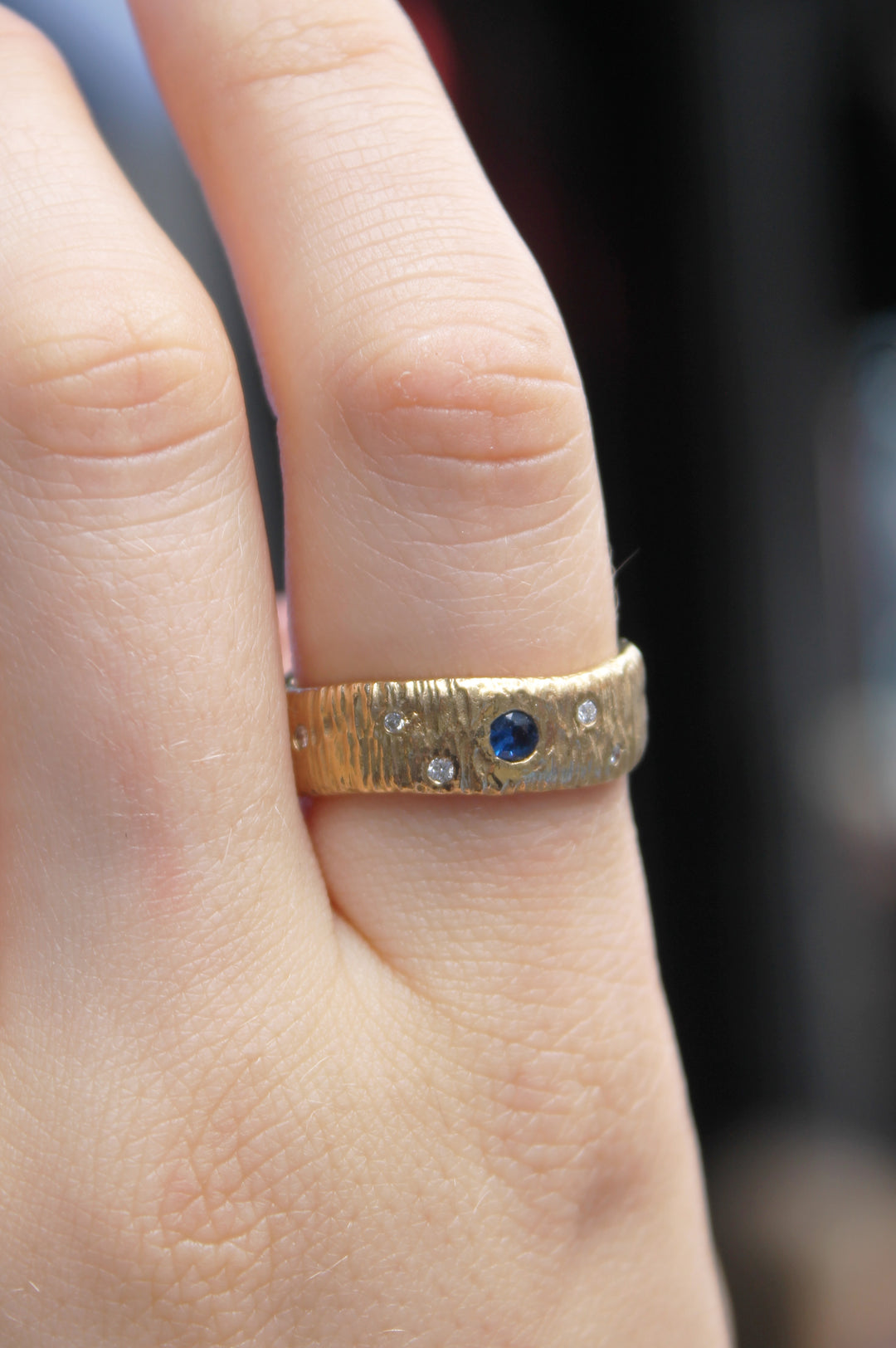 A hand wears the Etta Ring with Sapphire & Diamonds by Gallardo & Blaine Designs, featuring a 9ct yellow gold hammered band set with a central sapphire and two diamonds.