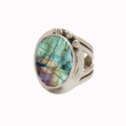 A silver statement cocktail ring featuring an oval-shaped labradorite stone with a spectrum of iridescent colors. The stone is set in a bezel setting, and the ring design includes three small round accent stones on one side of the band, enhanced by colourful gemstones for added brilliance. This is Eyetelia Ring in various gemstones by Gallardo & Blaine Designs.