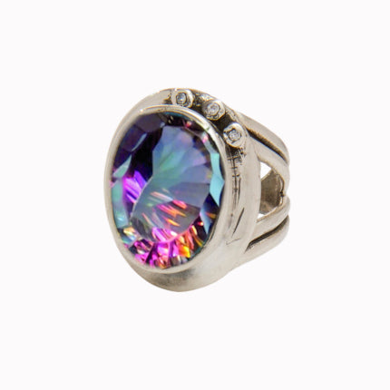 The Eyetelia Ring in various gemstones by Gallardo & Blaine Designs features a large, multicolored iridescent oval gemstone at its center. The wide band, designed with an adjustable design, splits into three parts near the top and is adorned with three small, clear stones along the edge of the main gemstone setting.