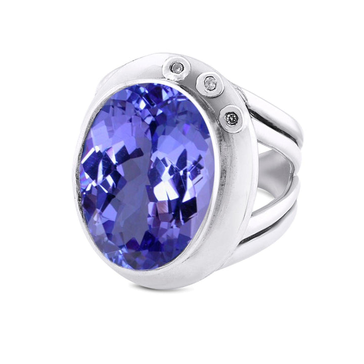 A silver statement cocktail ring featuring a large, oval-shaped purple gemstone set in the center. The band has a unique, open design with two thin strips on each side. Additionally, this ring is adorned with three small round diamonds on the side of the main gemstone. Presenting Gallardo & Blaine Designs' Eyetelia Ring in various gemstones.