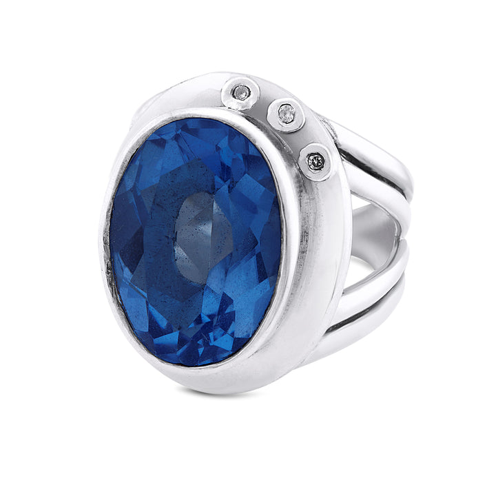 A stunning statement cocktail ring, the Eyetelia Ring in various gemstones by Gallardo & Blaine Designs features a large oval blue gemstone at its center, flanked by three small round white gemstones on one side. The band has a split shank style and an adjustable design. The faceted blue gemstone reflects light brilliantly, making it truly eye-catching.