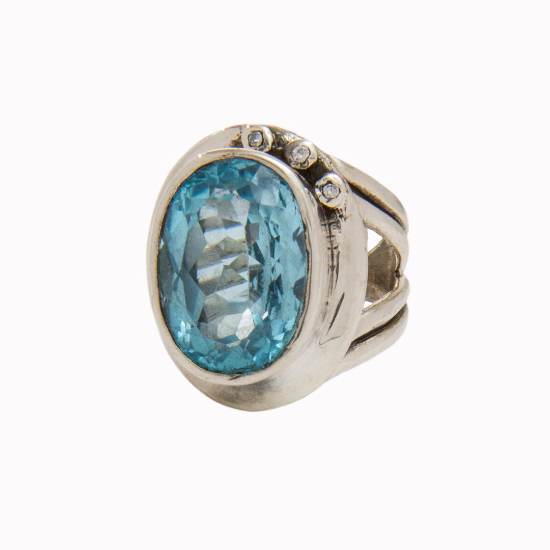 The Eyetelia Ring in various gemstones by Gallardo & Blaine Designs features a large oval-shaped blue gemstone set in the center. The faceted gemstone enhances its sparkle, while the band is adorned with three small round diamonds on one side, making it perfect for those who love colourful gemstones.