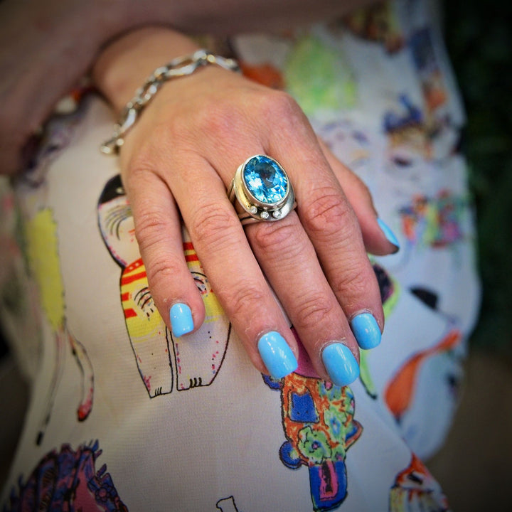 A close-up of a hand with blue nail polish, adorned with a statement Eyetelia Ring in various gemstones by Gallardo & Blaine Designs on the middle finger. The person is dressed in colourful, cat-patterned fabric, and an adjustable design silver bracelet graces the wrist.