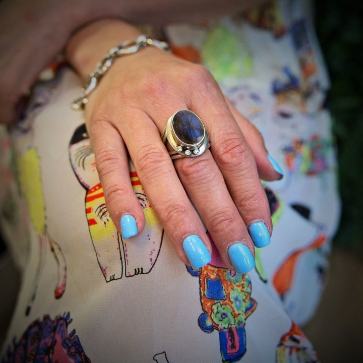 A hand rests on a fabric with colorful, illustrated cat designs. The nails are painted light blue, and the hand wears a large silver Eyetelia Ring in various gemstones by Gallardo & Blaine Designs. The person also wears a silver bracelet.