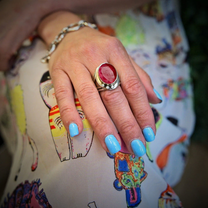 A hand with light blue nail polish rests gently on a colorful, cat-themed fabric. Adorned with the Gallardo & Blaine Designs Eyetelia Ring in various gemstones featuring a large, red stone and colourful gemstones, the hand draws attention. The background is slightly blurred, emphasizing the striking ring and polished nails.