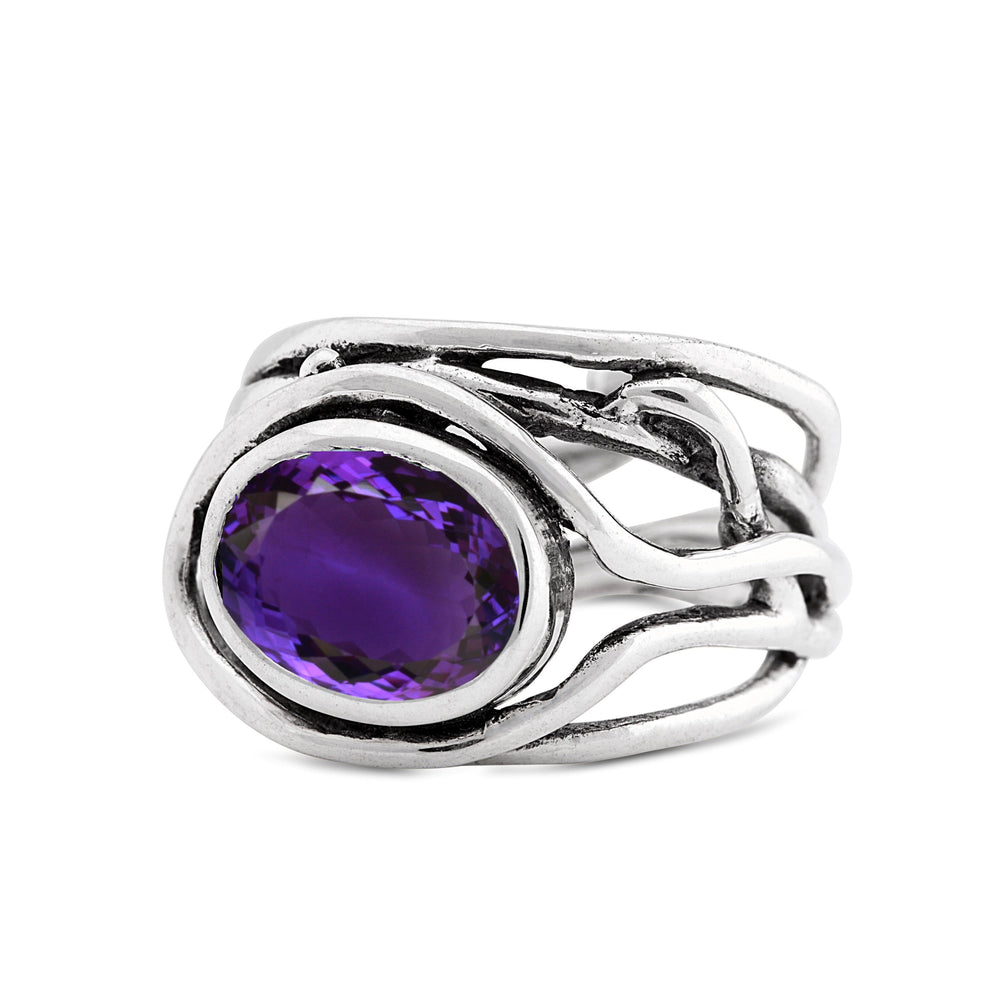 A Fossil Ring in silver & various gemstones by Gallardo & Blaine Designs with an intricate design featuring a large oval purple gemstone in the center. The band is composed of several intertwining silver strands, creating a unique and elegant look. This exquisite gemstone ring stands out as a stunning piece of jewelry.