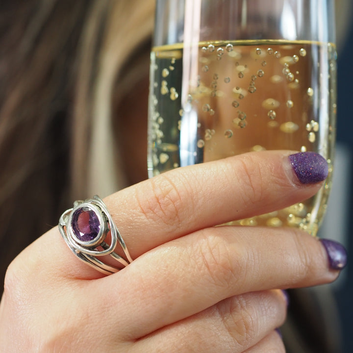 A hand with purple glittery nail polish holds a champagne flute filled with bubbly drink. The hand sports a Gallardo & Blaine Designs Fossil Ring in silver & various gemstones with a large, oval purple gemstone. The background is blurred and out of focus.