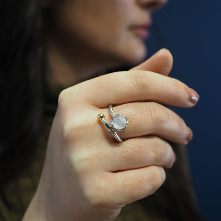 Close-up of a person holding their hand near their face, showcasing an elegant adjustable Honeysuckle Ring in Silver Gold & gemstones. The silver and gold jewelry from Gallardo & Blaine Designs features a round, translucent stone set in a simple band design with a small gold ball accent. The person is in partial profile, against a dark background.